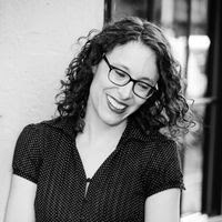 Black and white photo of a person with a dark, short-sleeved collared shirt, medium length curly hair, and dark rimmed glasses. Her face is tilted to the side with a large smile. Behind her looks like a window on the right-hand side.